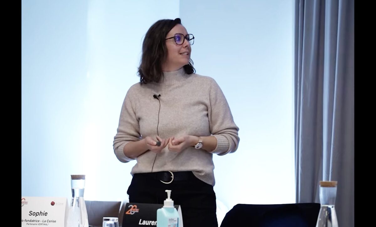 Femme parlant lors d'une conférence, portant des lunettes et un micro.